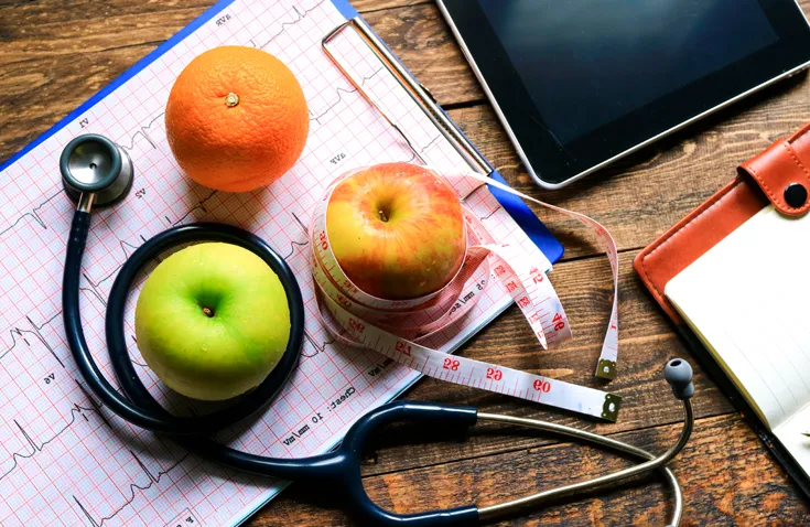 Apples and oranges wrapped around stethoscope and measuring tape on wooden desk.